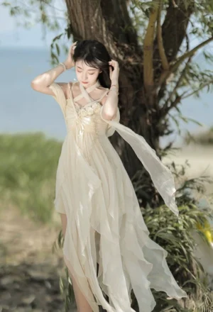 Cream halter JSK jumper skirt with asymmetric irregular hem - front three-quarter view outdoor seaside setting showing cross halter neckline and flowy chiffon sash details