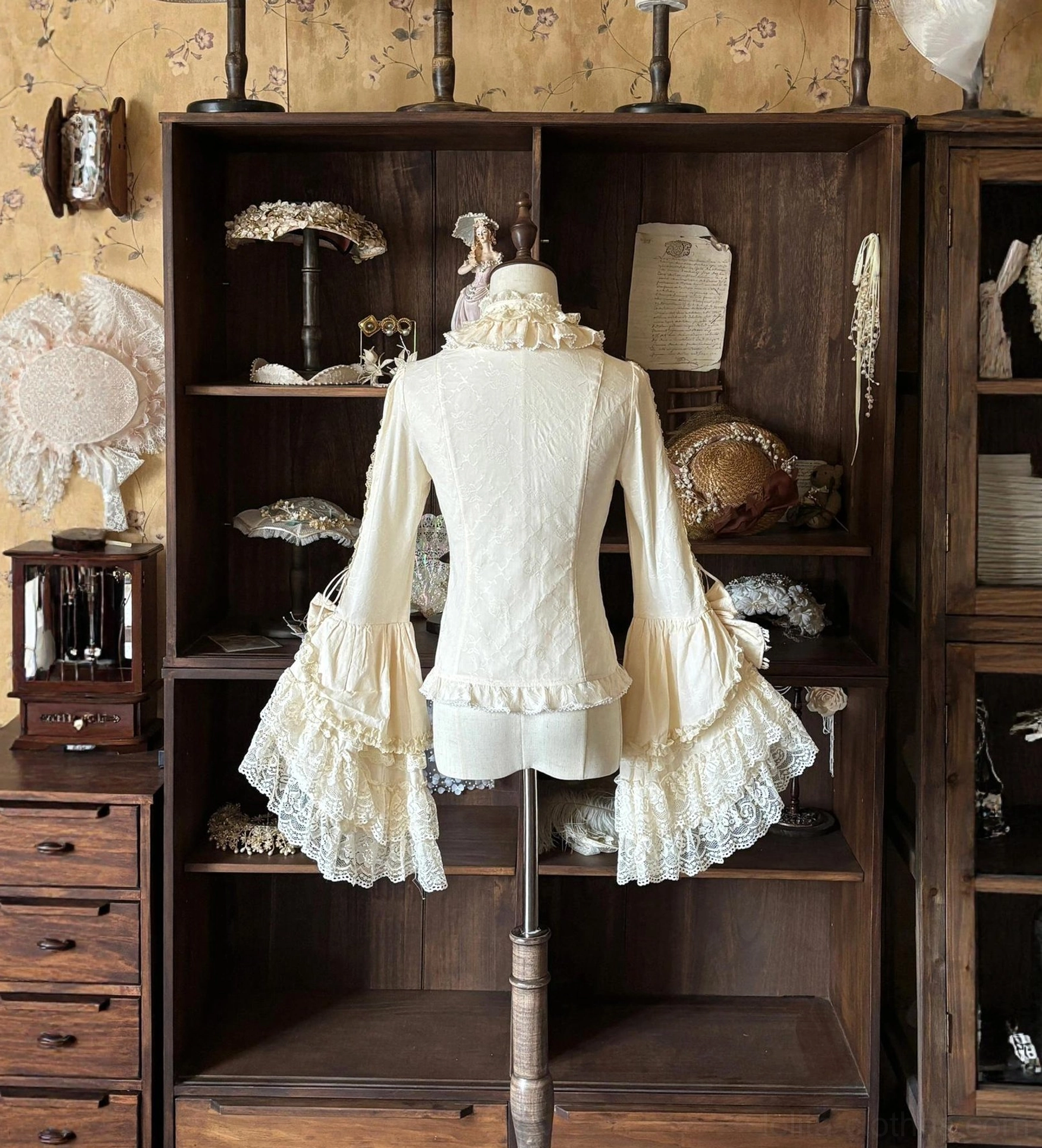 Cream off-white lolita blouse with fitted silhouette and dramatically flared lace bell sleeves shown from back three-quarter view on vintage display cabinet