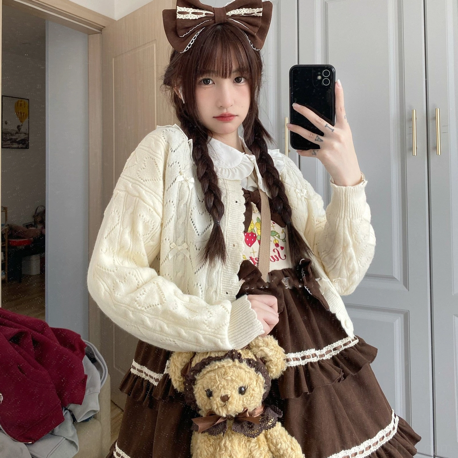 Front mirror selfie of cream hollow out Lolita cardigan featuring cable knit texture, button closure, and bow details, worn with twin braids and bow hair accessory