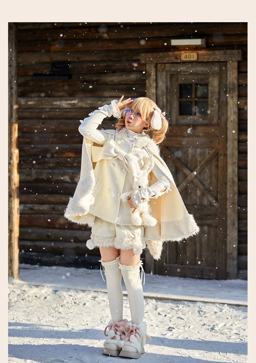 Cream colored winter lolita cape and shorts set with hooded design featuring ruffled short shorts and knee-high socks with pink fuzzy snow boots in snowy outdoor wooden cabin setting