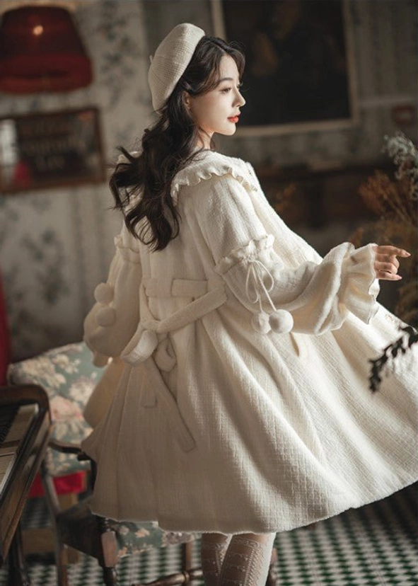 Side-back view of cream lolita winter coat showcasing A-line silhouette, back waist bow belt, drawstring waist, and pom-pom sleeve pendants on checkered floor