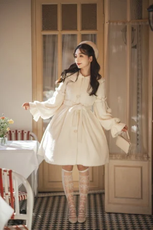 Cream puff winter lolita coat with heart-shaped buttons and removable pom-pom trims worn by model in vintage dining room setting, featuring round lapel collar and fluffy gold sable fabric