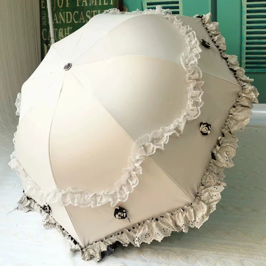 Cream white foldable lolita parasol with elegant double-layer ruffled lace trim and decorative black metal fasteners, featuring rounded dome canopy shape, displayed fully opened from rear top three-quarter view on tabletop with green window background