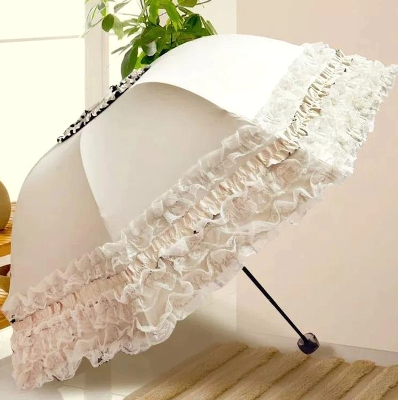 Cream white lolita lace ruffled parasol with double-layer ruffled lace trim, black beaded handle, shown from side three-quarter perspective in indoor home setting with wooden floor and green potted plant, partially folded display