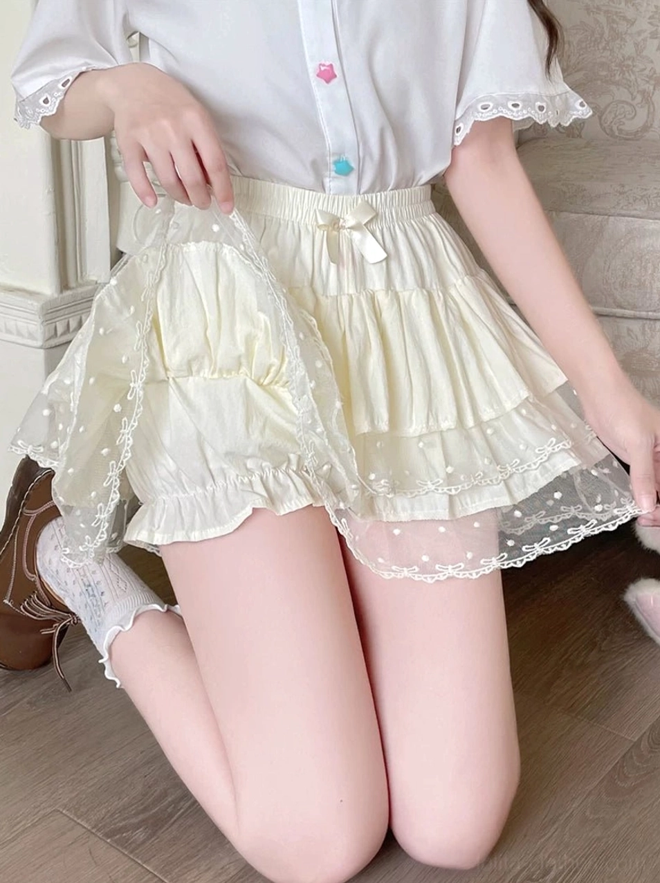 Cream white Lolita tiered bloomers side kneeling view in vintage bedroom background showcasing high elastic waist with decorative bow polka dot lace hem and multi-layer ruffle silhouette construction