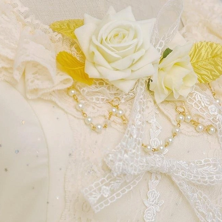 Close-up flat lay view of cream yellow Lolita collar accessory with large white artificial rose, gold leaf accents, pearl chain and lace bow decorations placed on cream sheer fabric