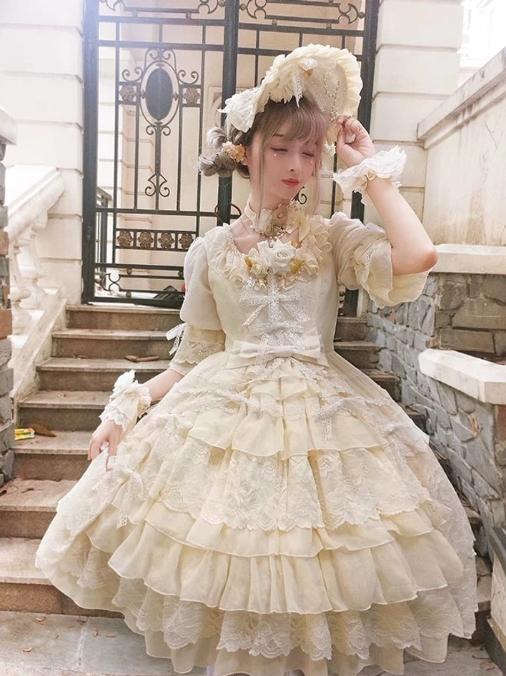 Cream yellow Sweet Lolita OP dress with lace-covered tiered skirt, floral brooch at neck, and puff half sleeves, front three-quarter villa stairs view