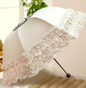 Creamy white lace ruffled parasol displayed in elegant interior setting with wooden flooring and greenery, showing layered ruffled lace trim and decorative jewel detail on canopy top