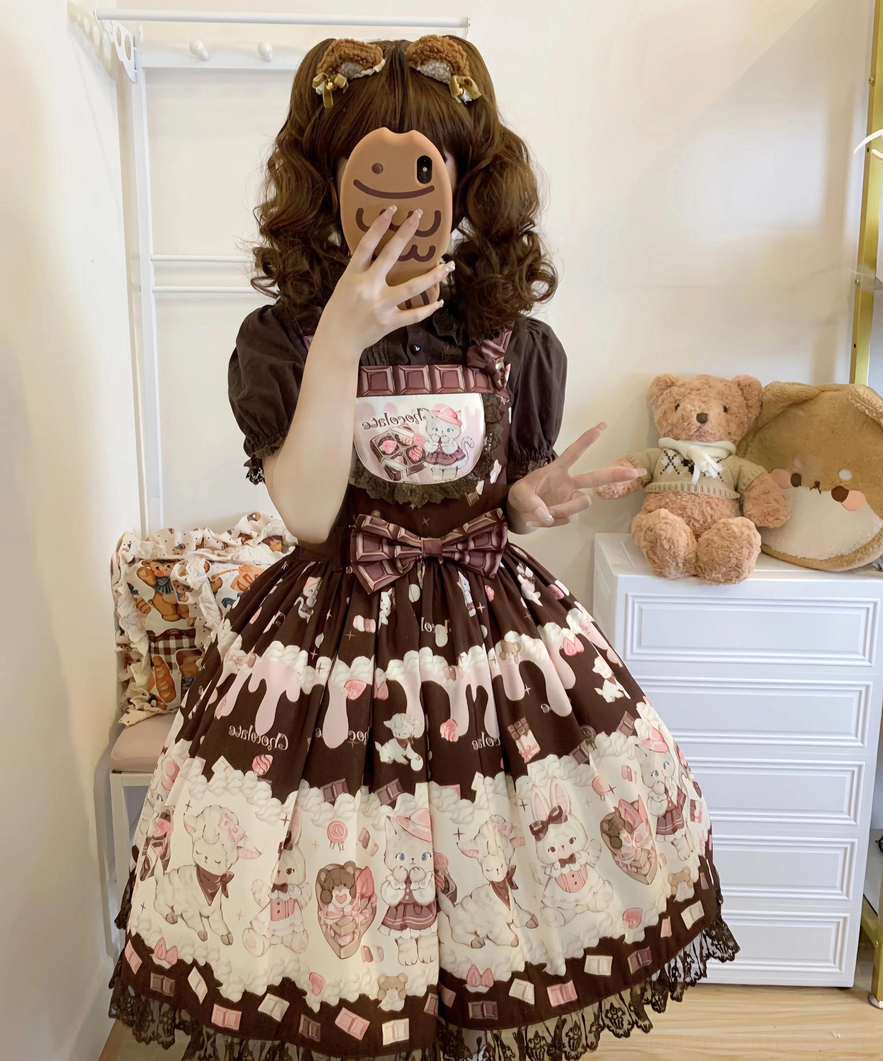 Brown curly-haired twin-tailed model taking hidden face selfie wearing dark brown and cream color-block Lolita salopette dress with bold two-tone print, large front chest bow, and layered printed skirt panels