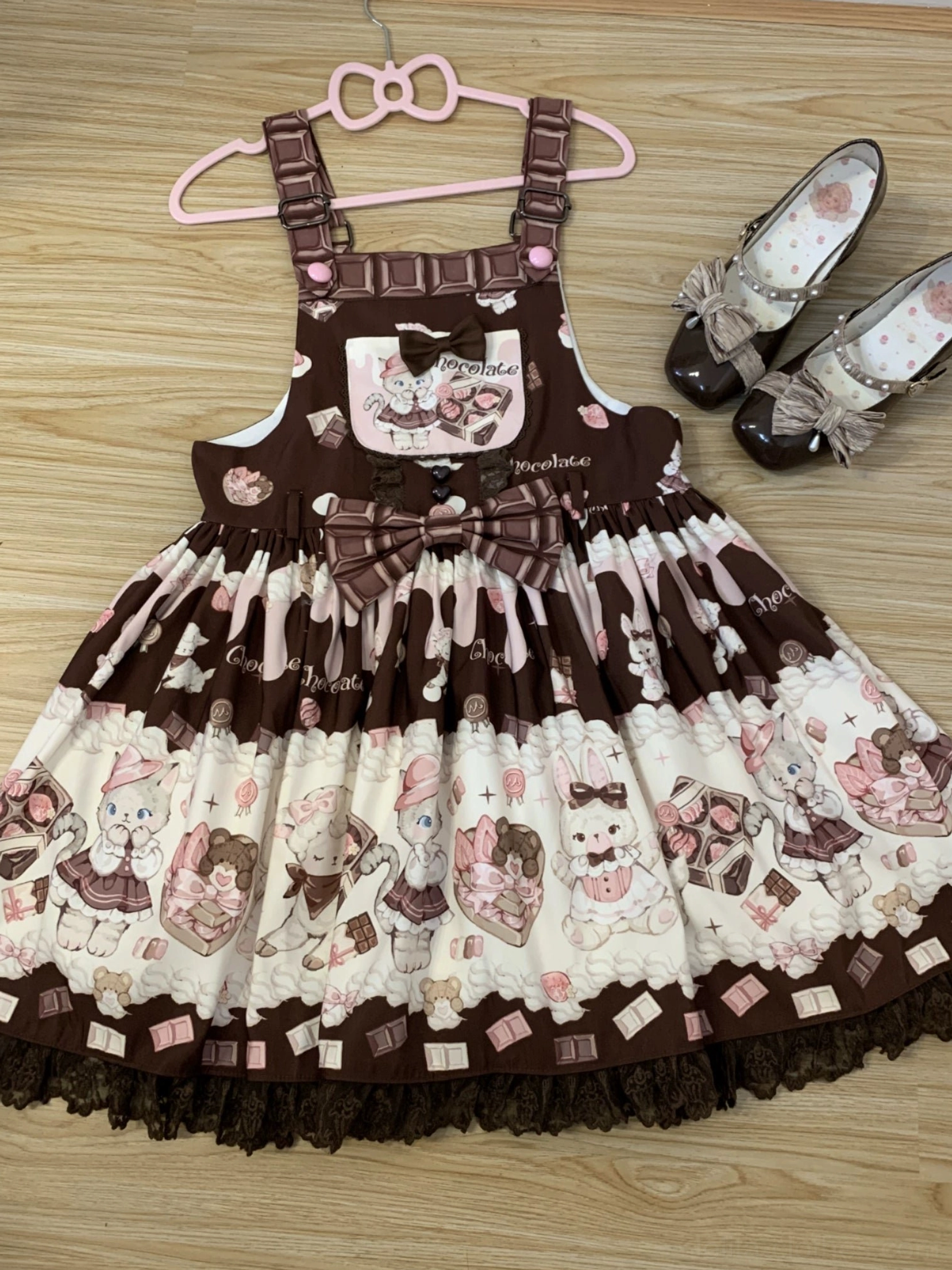 Dark brown and cream cat print Lolita salopette dress front view hanging display on wooden floor featuring layered printed panels, front waist bow decoration, lace skirt hem, and matching sweet Lolita Mary Jane shoes