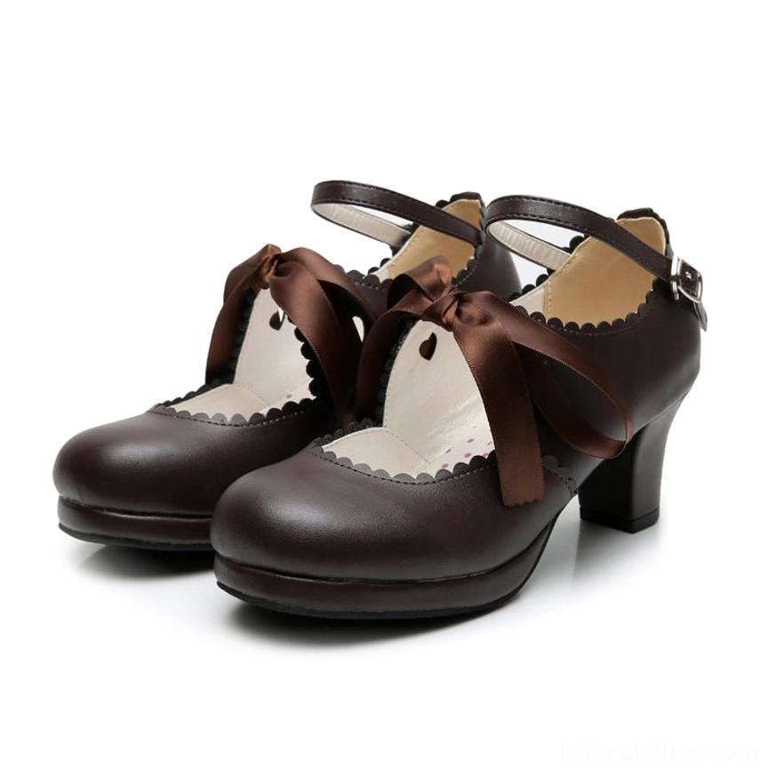 Dark brown handcrafted lolita high heel shoes flat lay front display showing scalloped collar, brown ribbon bow, ankle buckle strap, heart cutout inside vamp, and glossy brown leather on neutral background