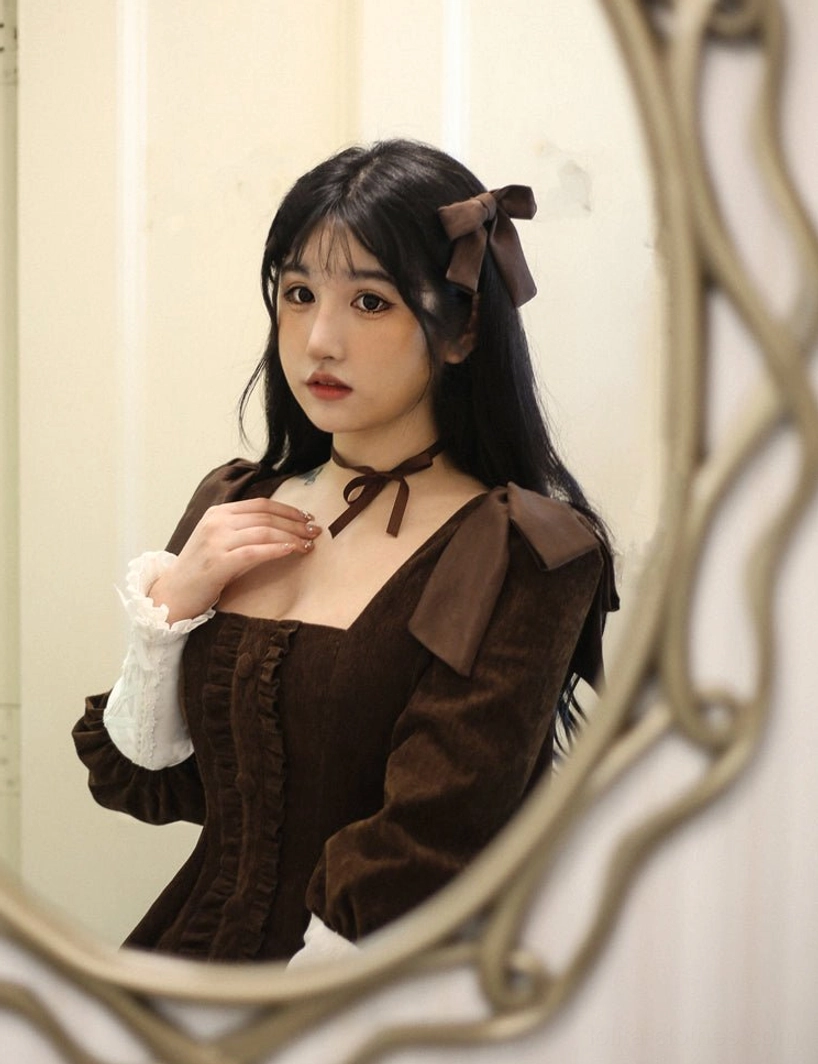Dark brown velvet Lolita OP dress close-up mirror portrait with simple indoor wall background, displaying shoulder bow decorations, neck strap bow, contrast white cuffs, and square neckline detail
