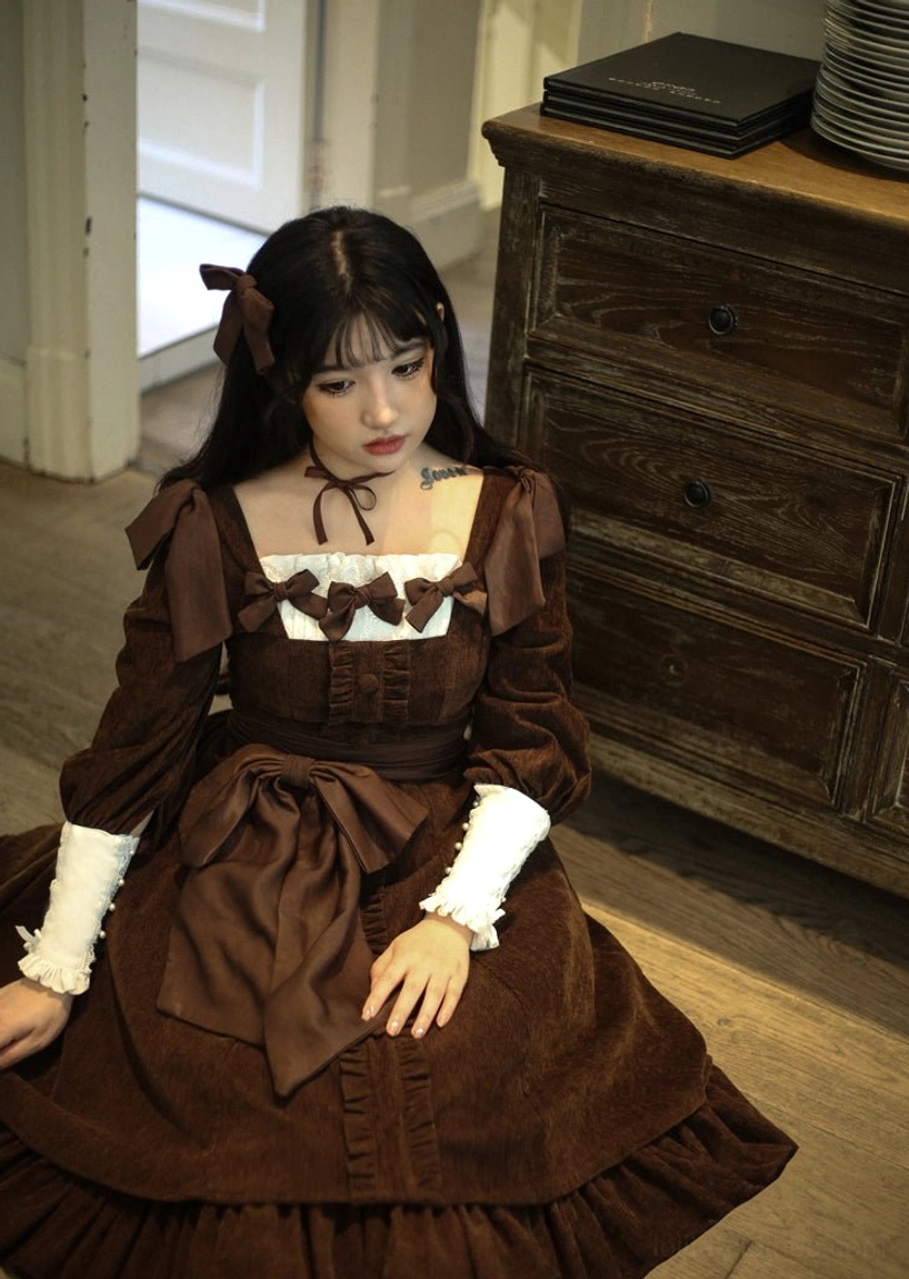 Dark brown velvet classic Lolita OP dress front full-body close portrait in vintage wooden furniture indoor space, displaying chest bow trim, large front waist bow, contrast white cuffs, and velvet ruffled skirt hem