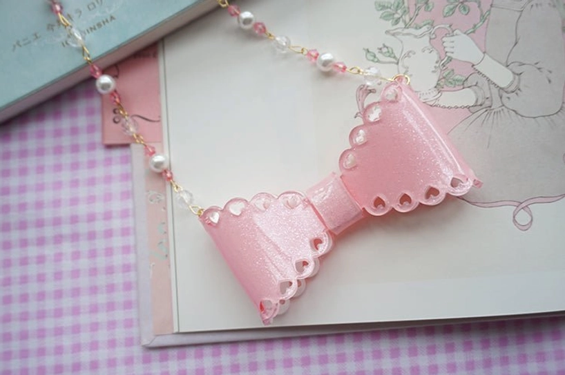 Dark pink glittery scalloped resin bow necklace with pearl beaded chain detail on vintage illustrated book background soft focus