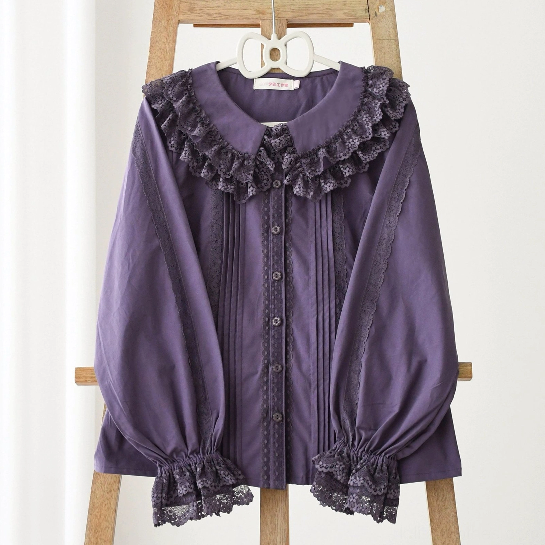 Close-up front flat display of dark purple solid Lolita long-sleeve blouse on wooden hanger with pure white background, showing lace ruffled Peter Pan collar, front pleated button placket, and lace-trimmed cuffs