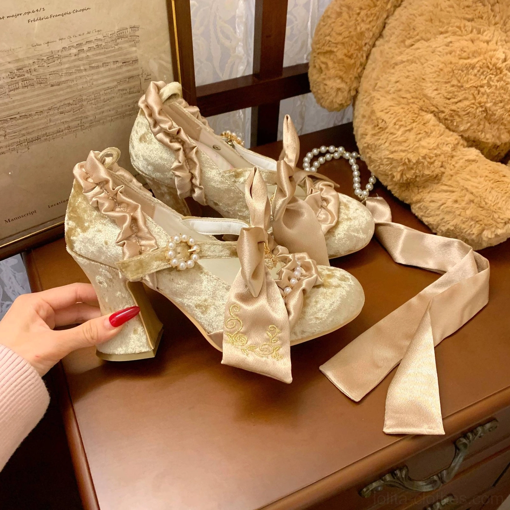Hand displaying champagne gold removable satin bow strap and pearl decorated suede-like embroidered Lolita heel on wooden dresser