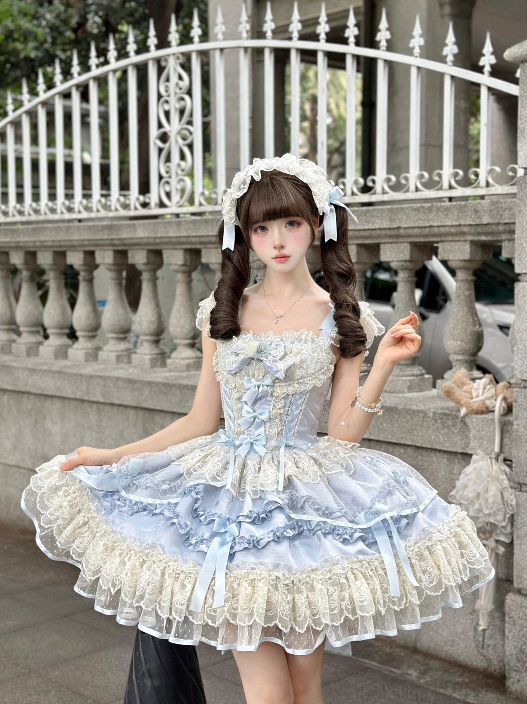 Blue ballet-style sweet lolita jumper skirt front view on outdoor vintage iron balcony, corset bodice, layered lace skirt, white headdress, model lifting skirt hem