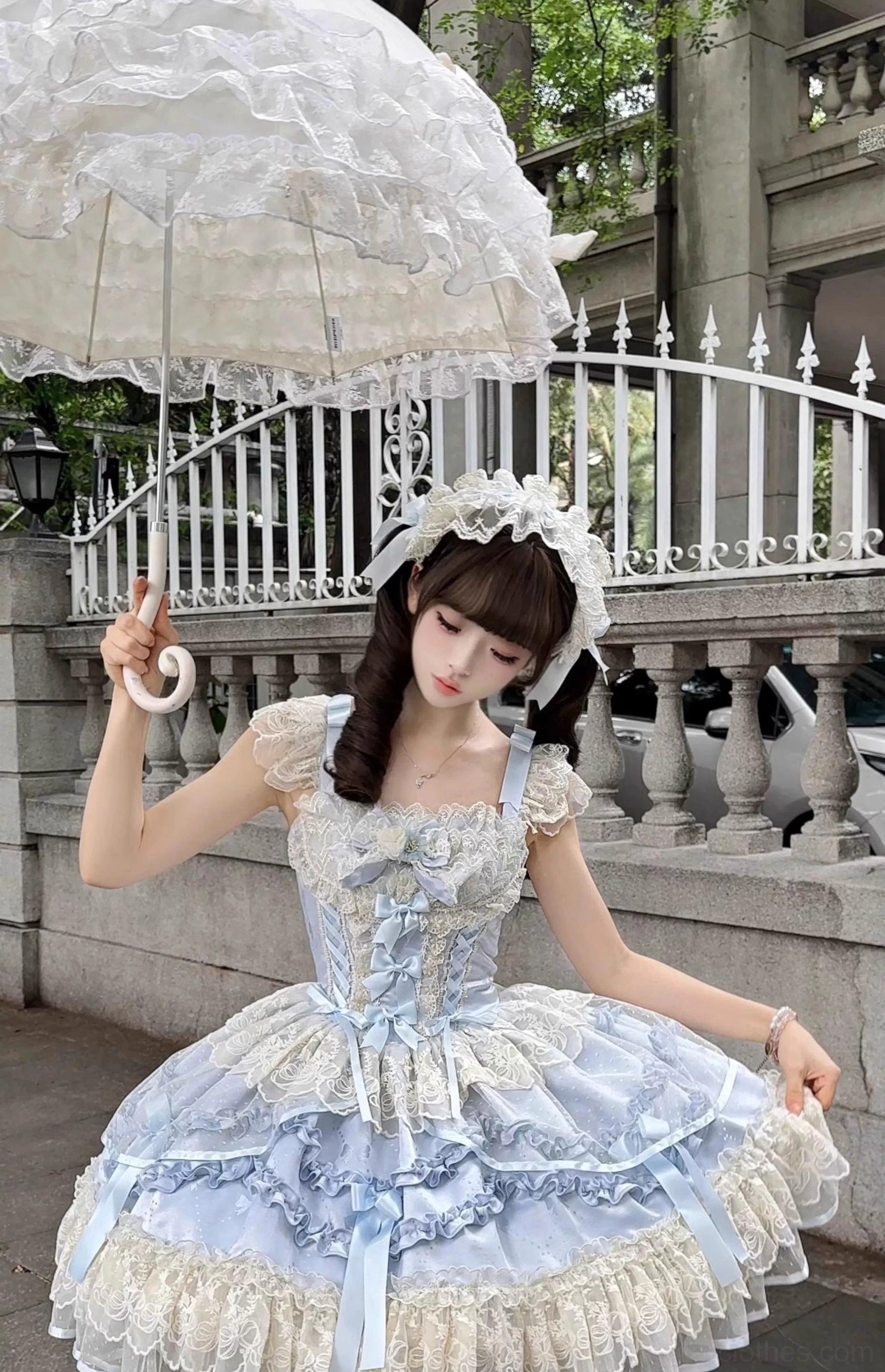 Blue ballet-style sweet lolita JSK dress front view outdoor vintage balcony with layered lace skirt, corset bodice, white headdress, model holding vintage lace parasol