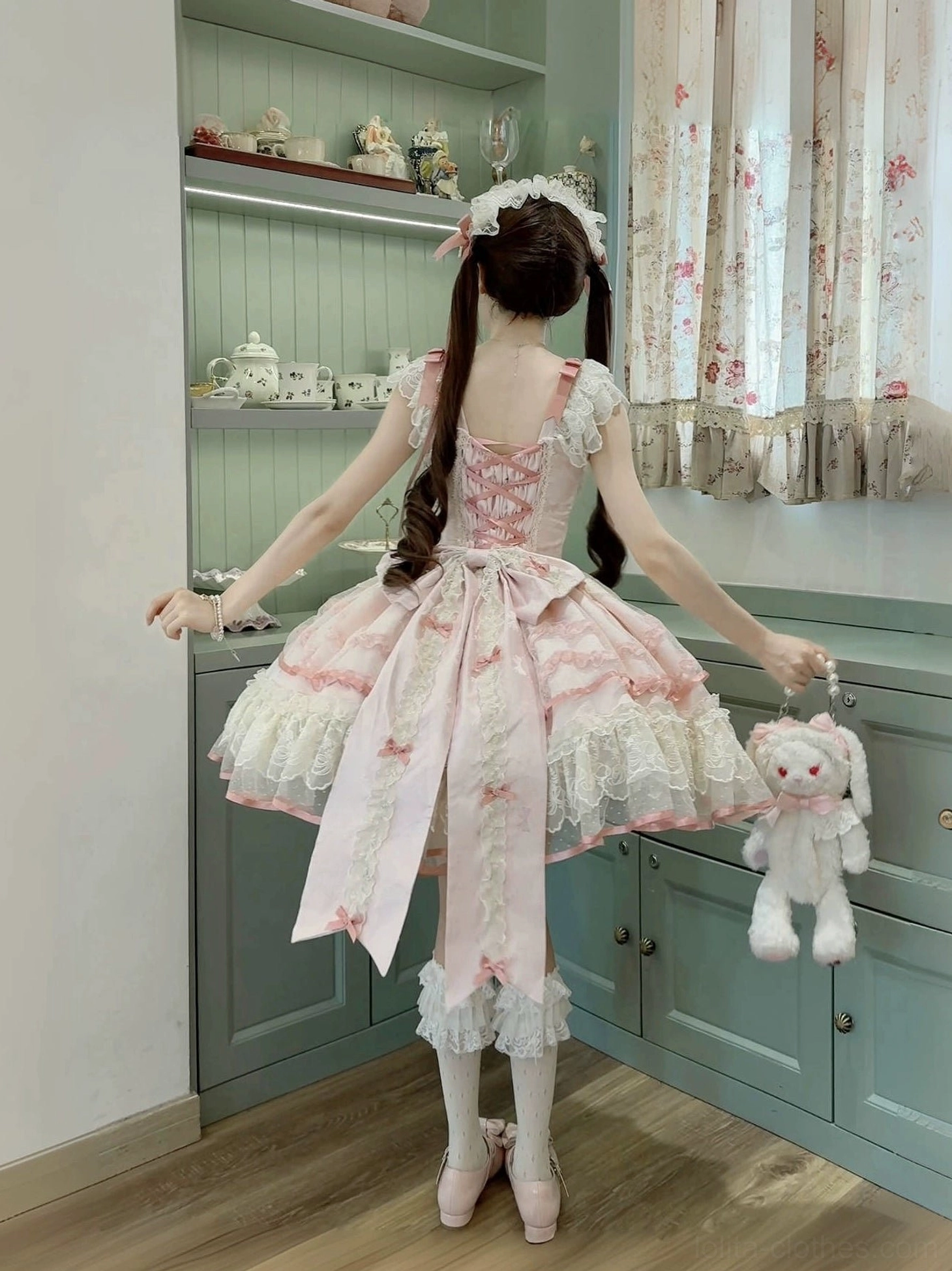 Pink Dreamy Butterfly JSK dress back view with large trailing bow, ruffled lace trim, white stockings, indoor vintage cabinet room styling with twin-tailed model holding plush rabbit