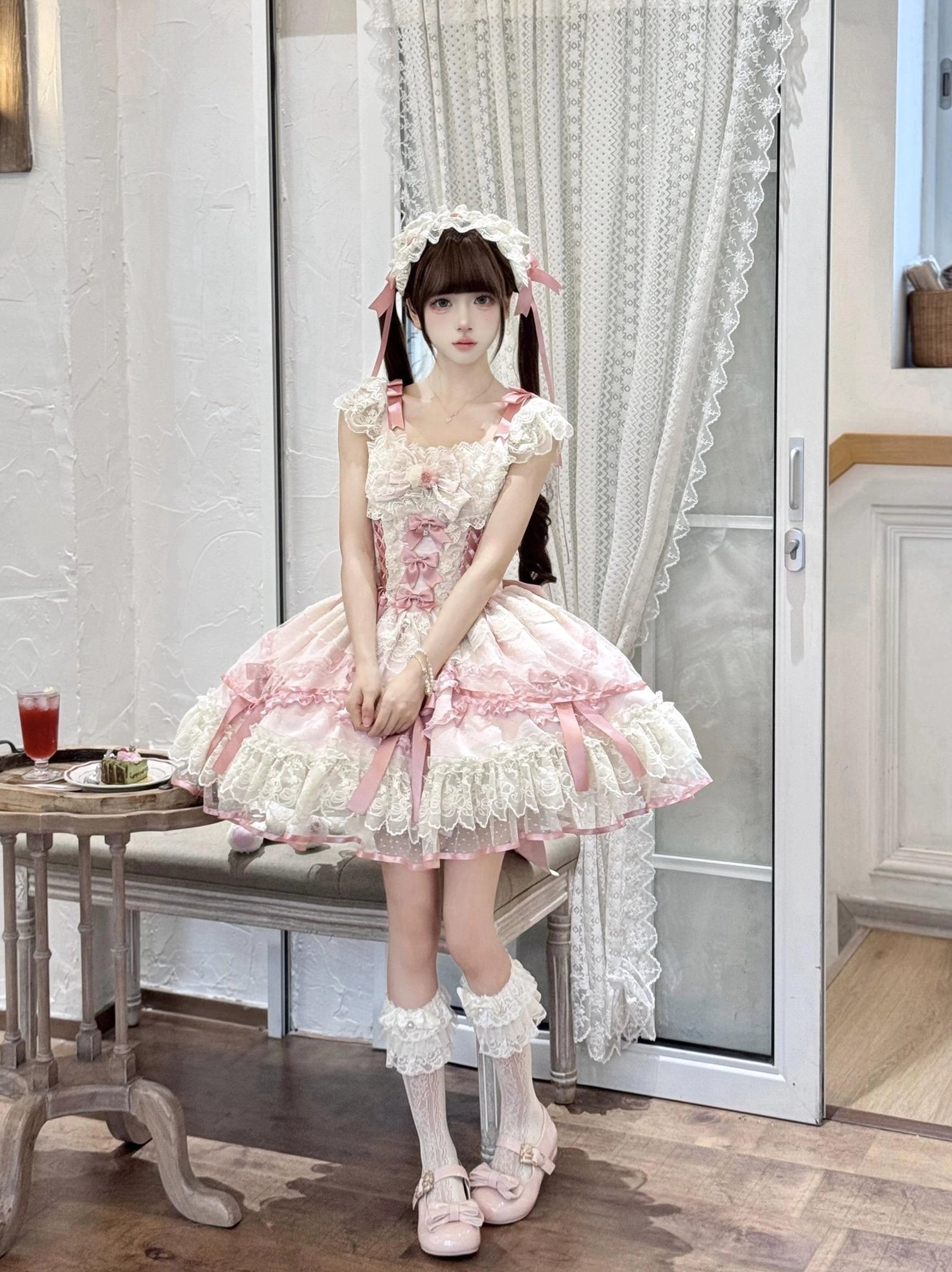 Pink sweet lolita jumper skirt front seated view with ruffled lace sleeves, matching hair headdress, white lace socks, model with folded hands pose, indoor tea studio