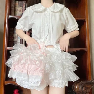 Dual-color lolita safety bloomers featuring white and pink organza options with cascading multi-layer ruffles, delicate lace leg hem, and elastic waistband styled under white doll collar blouse against vintage bookshelf background