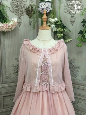 Dusty rose pink lolita cardigan with ruffled crew neckline and shoulders, featuring delicate lace-up front detail and dimensional ruffled waist trim, styled as part of a complete lolita coordinate with matching dress, displayed on dress form against vintage floral garden backdrop