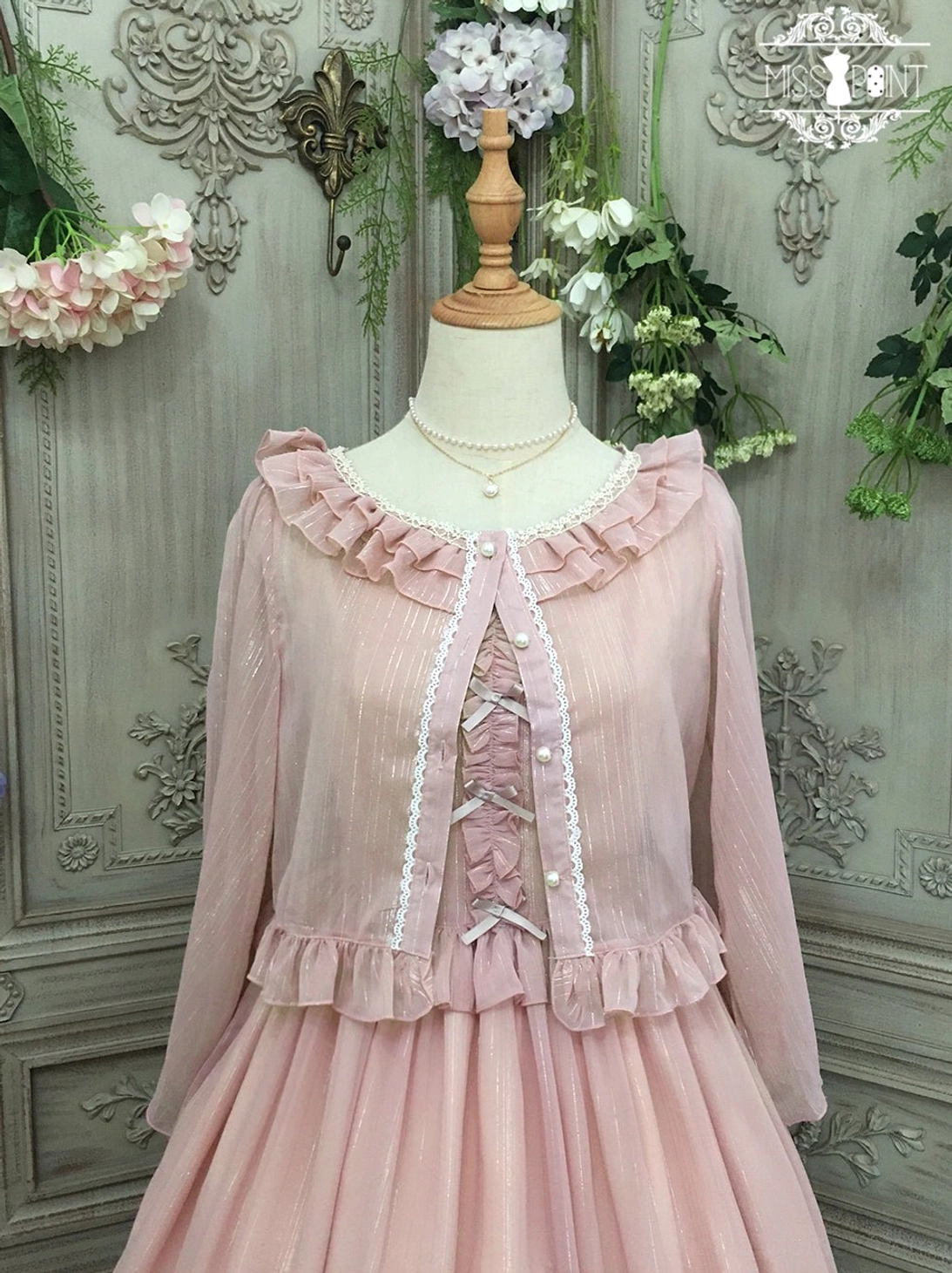 Dusty rose pink lolita cardigan with ruffled crew neckline and shoulders, featuring delicate lace-up front detail and dimensional ruffled waist trim, styled as part of a complete lolita coordinate with matching dress, displayed on dress form against vintage floral garden backdrop