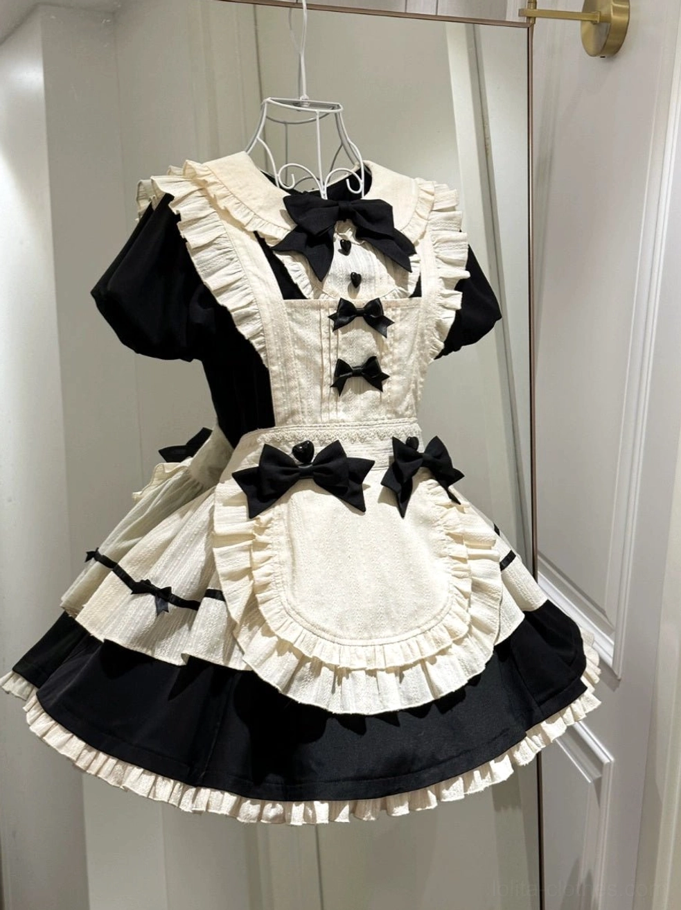 Short-sleeve halloween maid lolita apron OP dress on metal hanger indoor setting showing ruffled trims and multiple black bow decorations on bodice