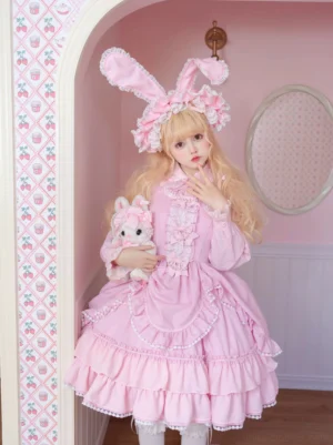 Pink long-sleeve classic lolita one-piece dress with ruffled bust detail and layered hemline held by model with plush rabbit toy indoor pink room