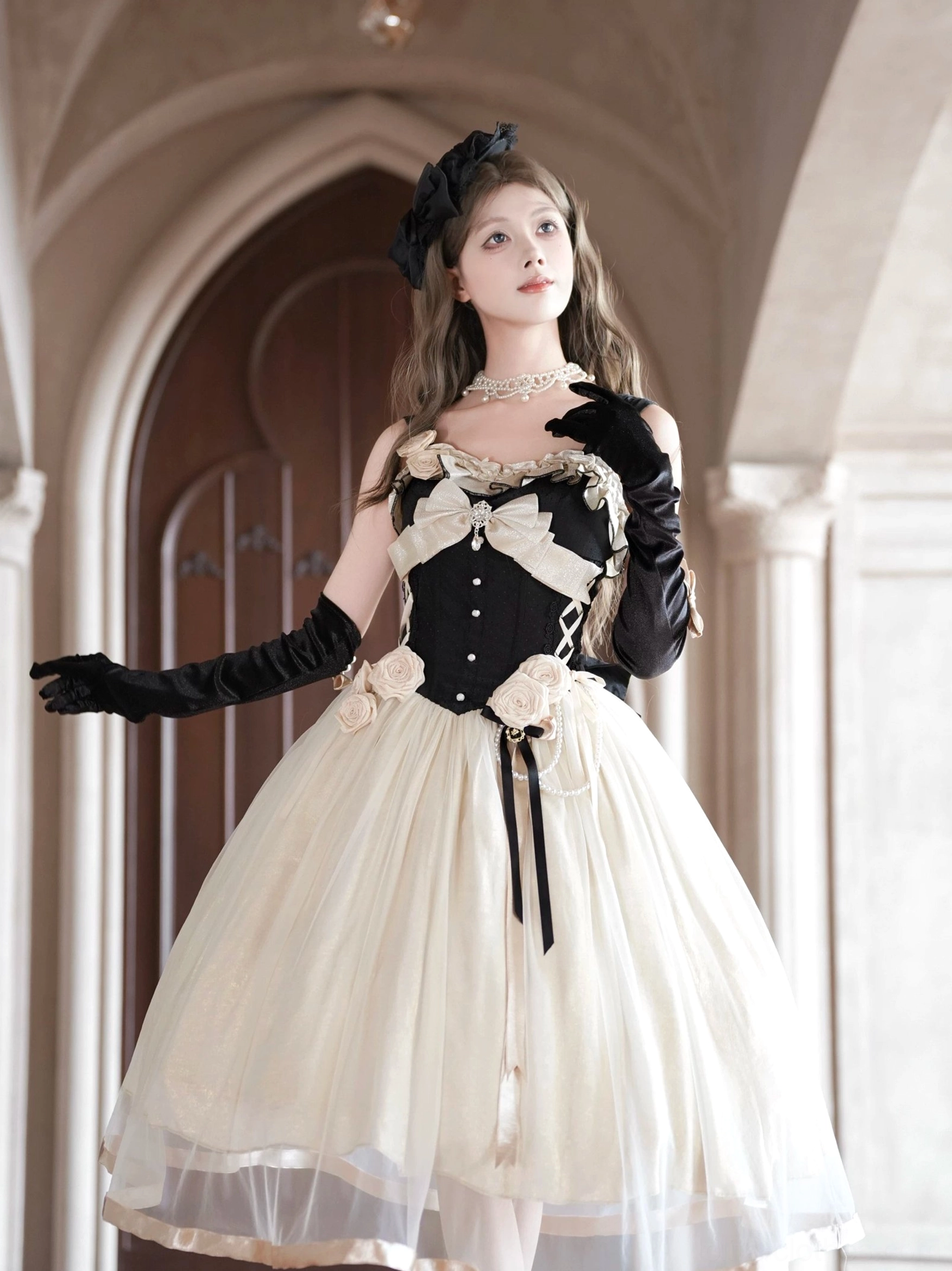 Front three-quarter angle of Classic Lolita jumper skirt in formal mansion hallway, showcasing large bust bow, 3D fabric waist roses, satin ribbon sash detail on skirt