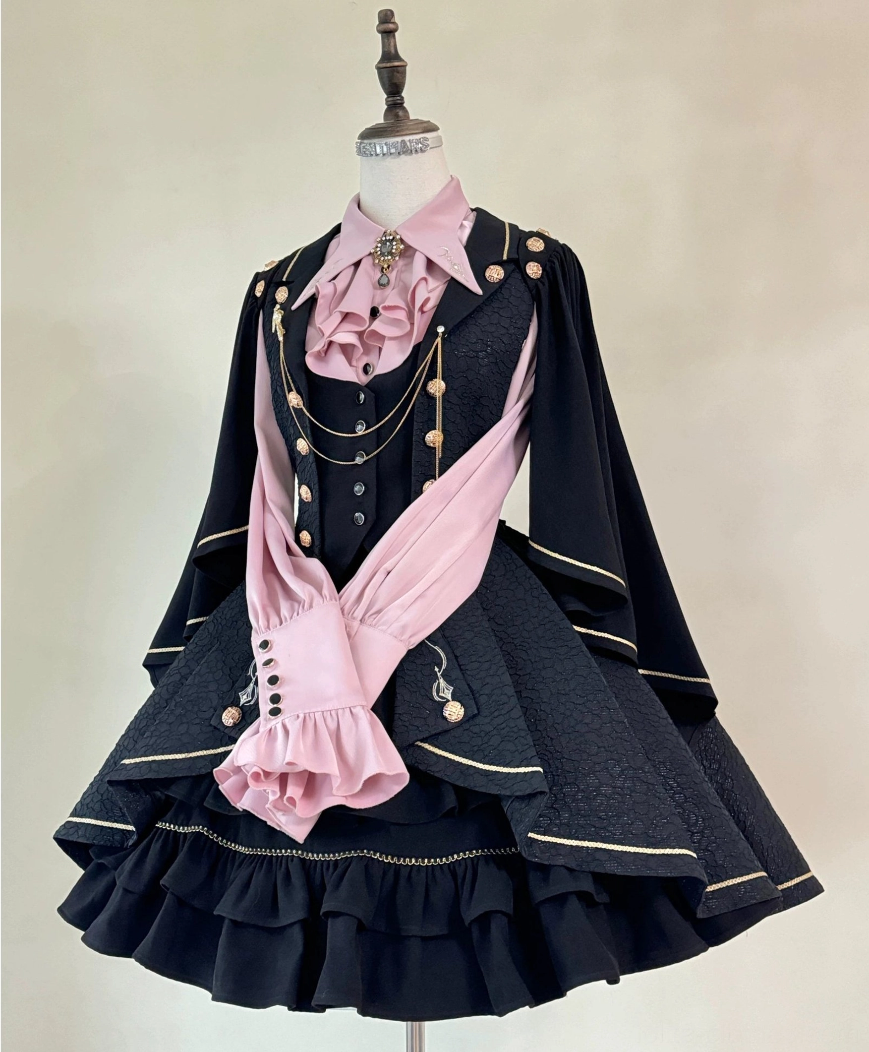 Fallen Moon Rule black and pink retro knight military lolita dress set, frontal static display on mannequin, light warm background, featuring pink ascot necktie, gold chain decorations, cape sleeves, and tiered ruffled skirt with gold trim