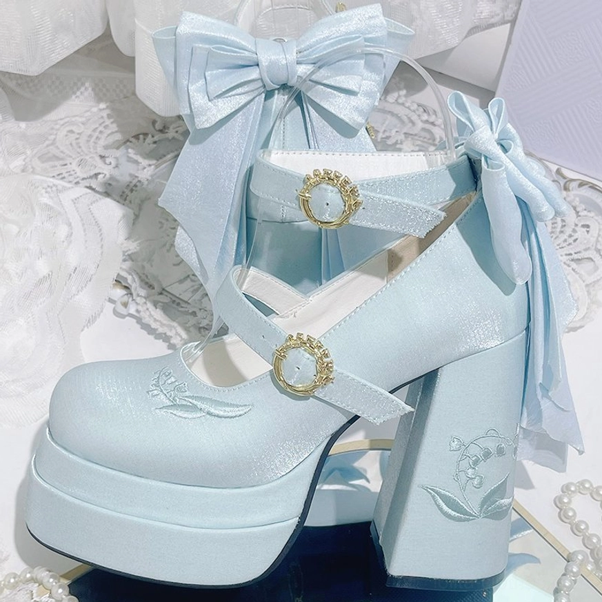 Baby blue satin lolita high heel shoes with detachable large satin back bow, gemstone buckles, and lily embroidery, three-quarter side flat lay view on sheer lace fabric backdrop