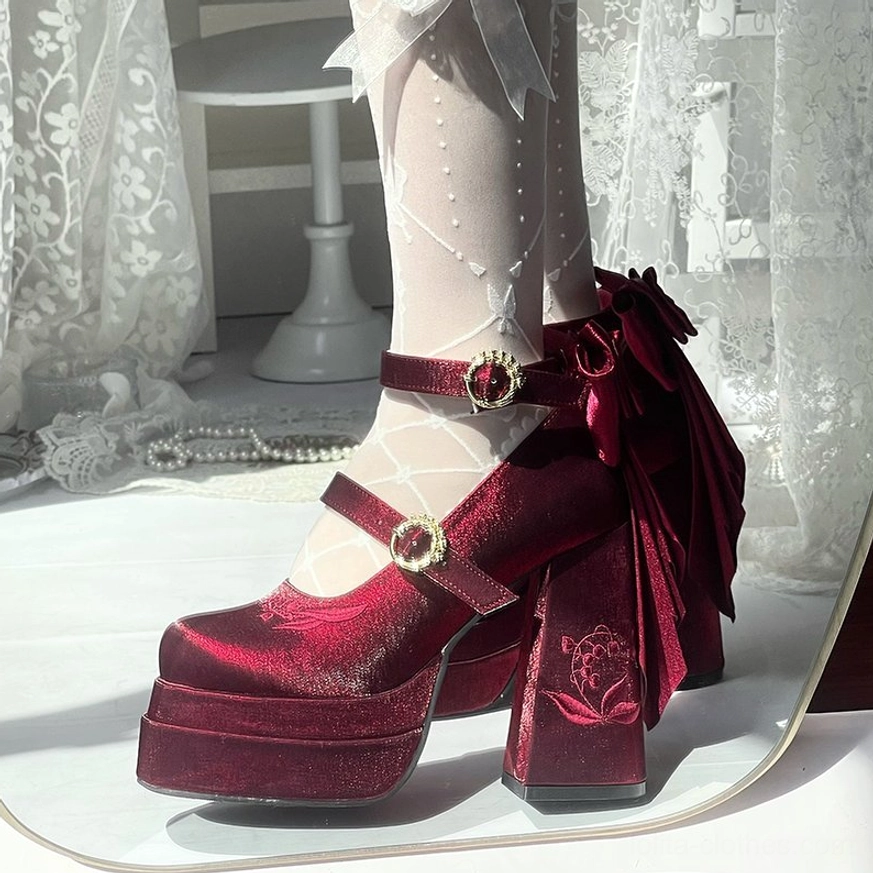 Burgundy velvet lolita high heel pumps with large velvet back bow, red gem straps, and floral embossed block heel, front side view worn with dotted white sheer stockings
