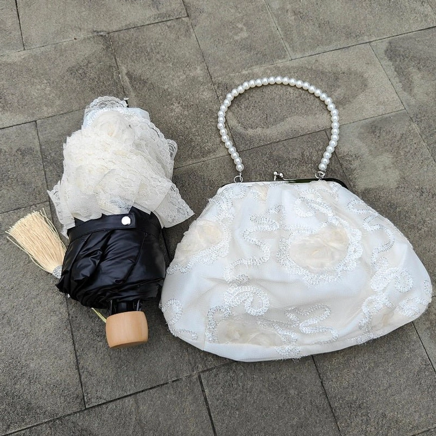 Compact folded lolita parasol paired with matching white lace handbag on stone pavement, showing portability and coordinated lolita accessories with wooden handle and pearl-topped handbag