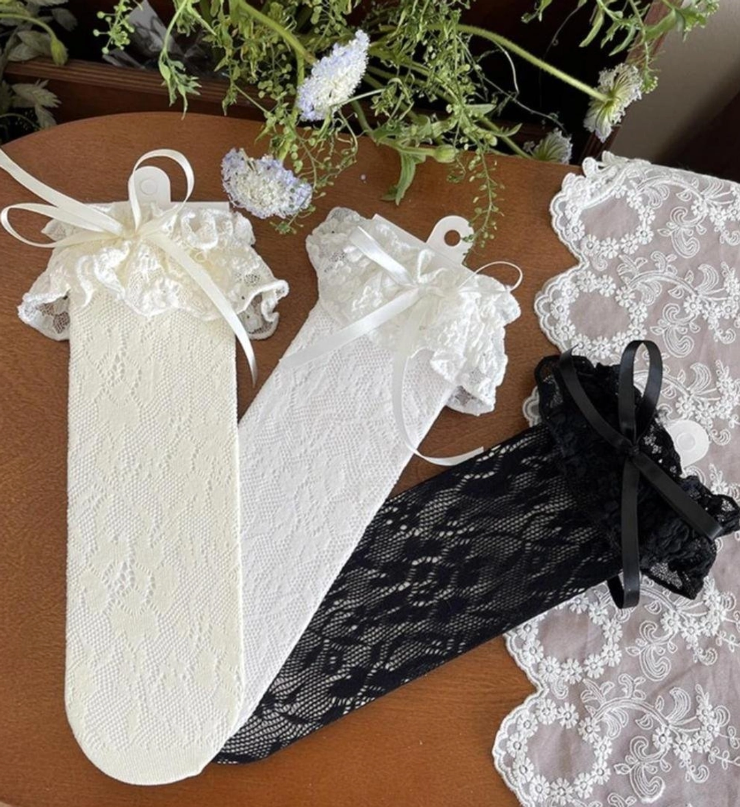 Complete collection of French Lolita mid-calf lace socks showing all three colorways - off-white pairs with white satin bows and black pair with black bow, flat lay display on wooden surface with decorative lace placemat