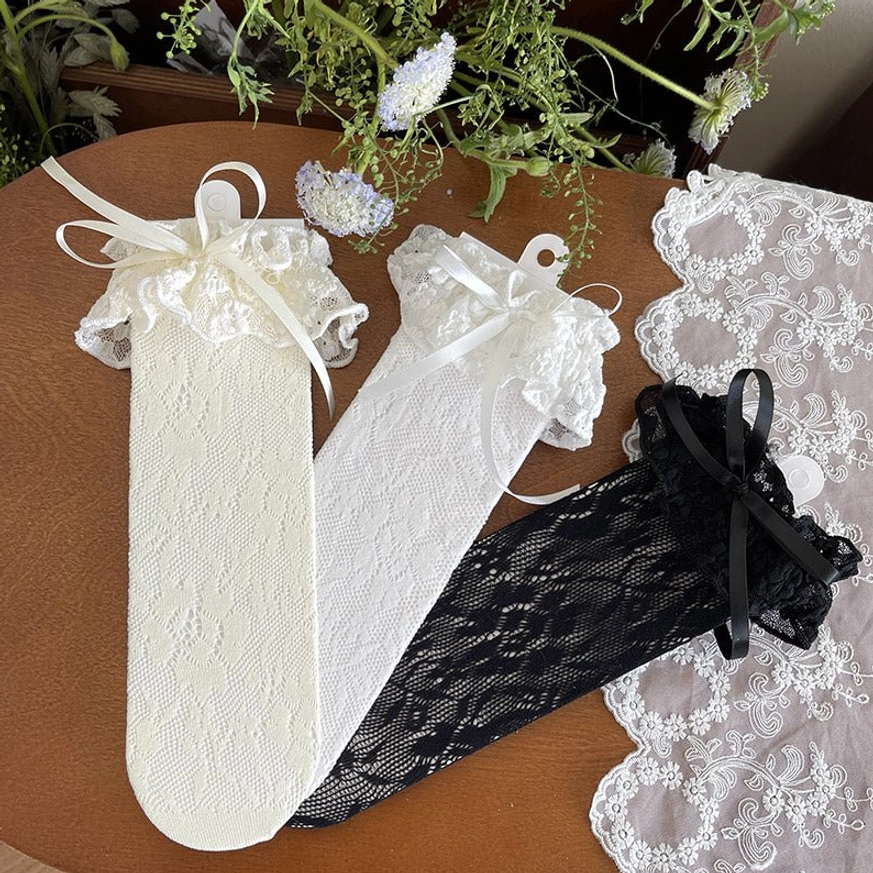 Full set French Lolita mid-calf lace socks overhead flat lay showing two off-white pairs with white bows and one black pair with black bow, all with ruffled cuffs on wooden table with plants