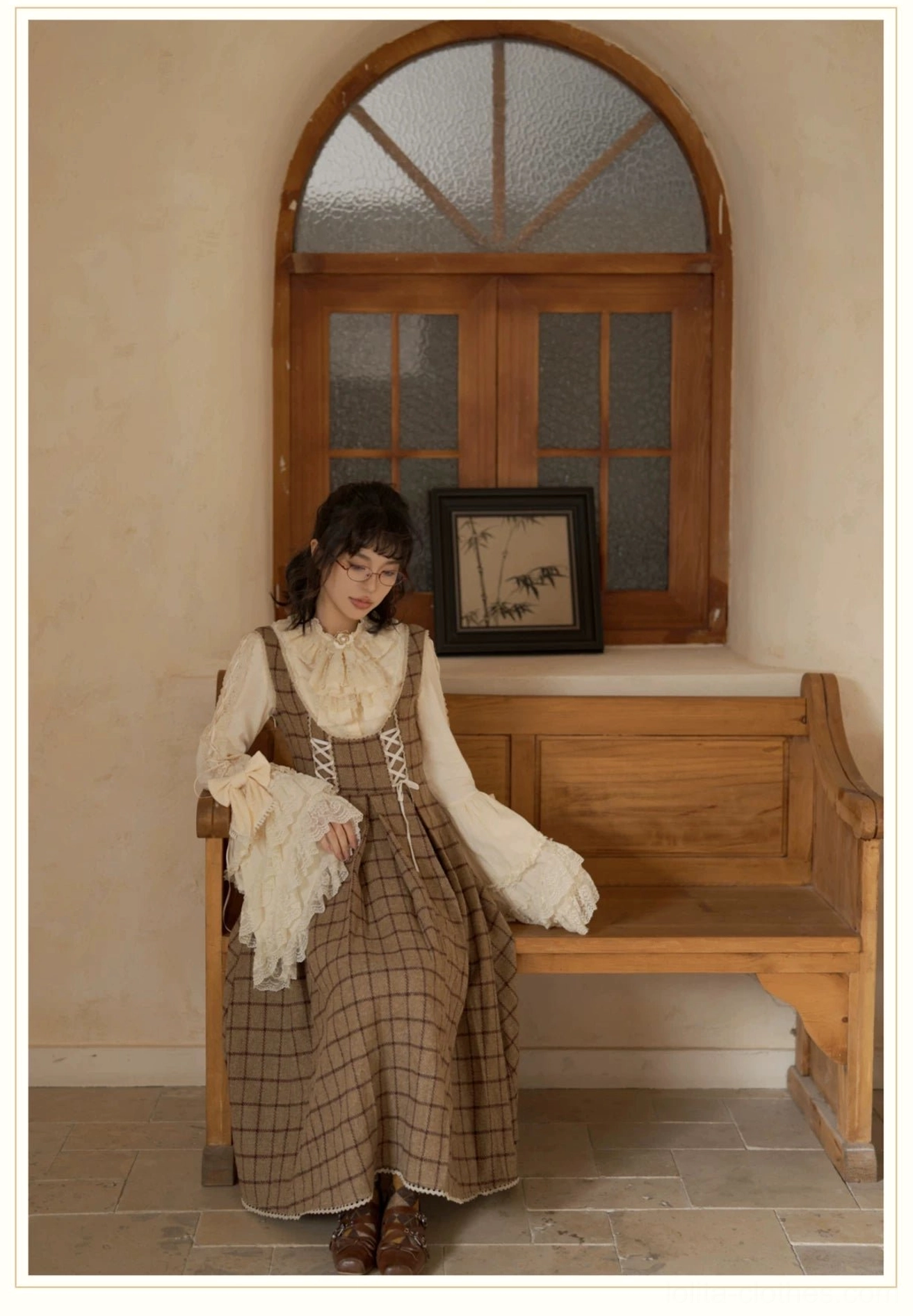 Full body lolita fashion coordination of cream hime sleeve blouse and plaid overall dress worn by model in vintage cottage setting with wooden window and bench holding lace scarf
