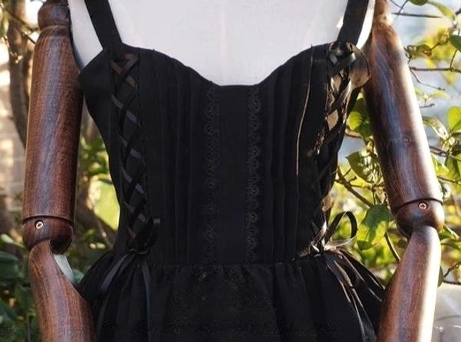 Garden Waltz black lolita JSK close-up detail shot of bodice lace-up ribbon front with delicate lace edge trim spaghetti straps vertical pleated structure fitted silhouette side adjustable ribbon details outdoor green plant background