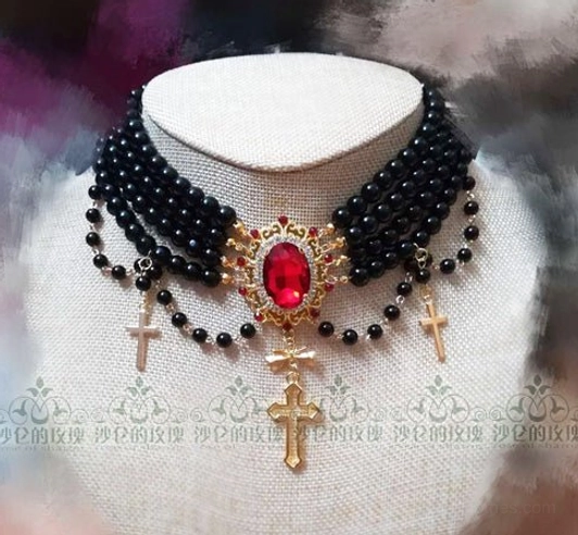 Gothic style black pearl tiered choker necklace with large red central gemstone and ornate gold cross pendants on white fabric display mannequin, featuring tiny black bead accents and dramatic dark artistic background