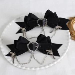 Pair of 3D bow Gothic Lolita hair clips with black satin bows, heart-shaped metal buckle, and chain pendants featuring tiny gun and lock charms, presented on white porcelain plate with golden trim