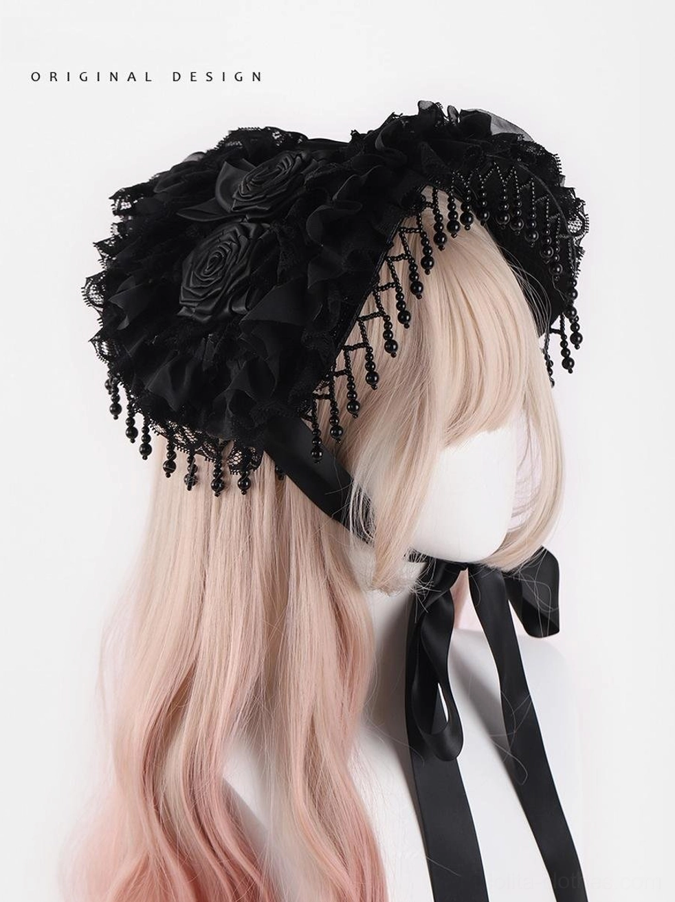 Gothic Lolita black bonnet hat with hand-knotted beaded tassel trim and handcrafted fabric roses displayed on mannequin with gradient pink-blonde curly wig, three-quarter front view showing black satin neck ties and ruffled lace base