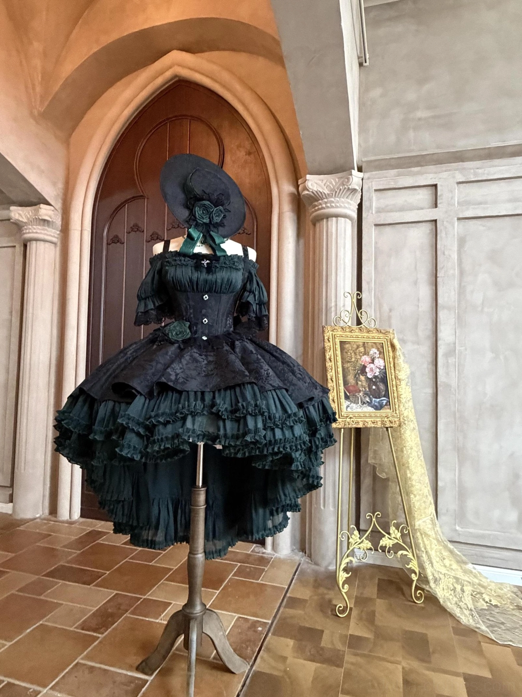 Dark teal Gothic Lolita boned JSK dress worn by model in European arched hallway with corset bodice, high-low layered ruffled skirt and formal matching hat