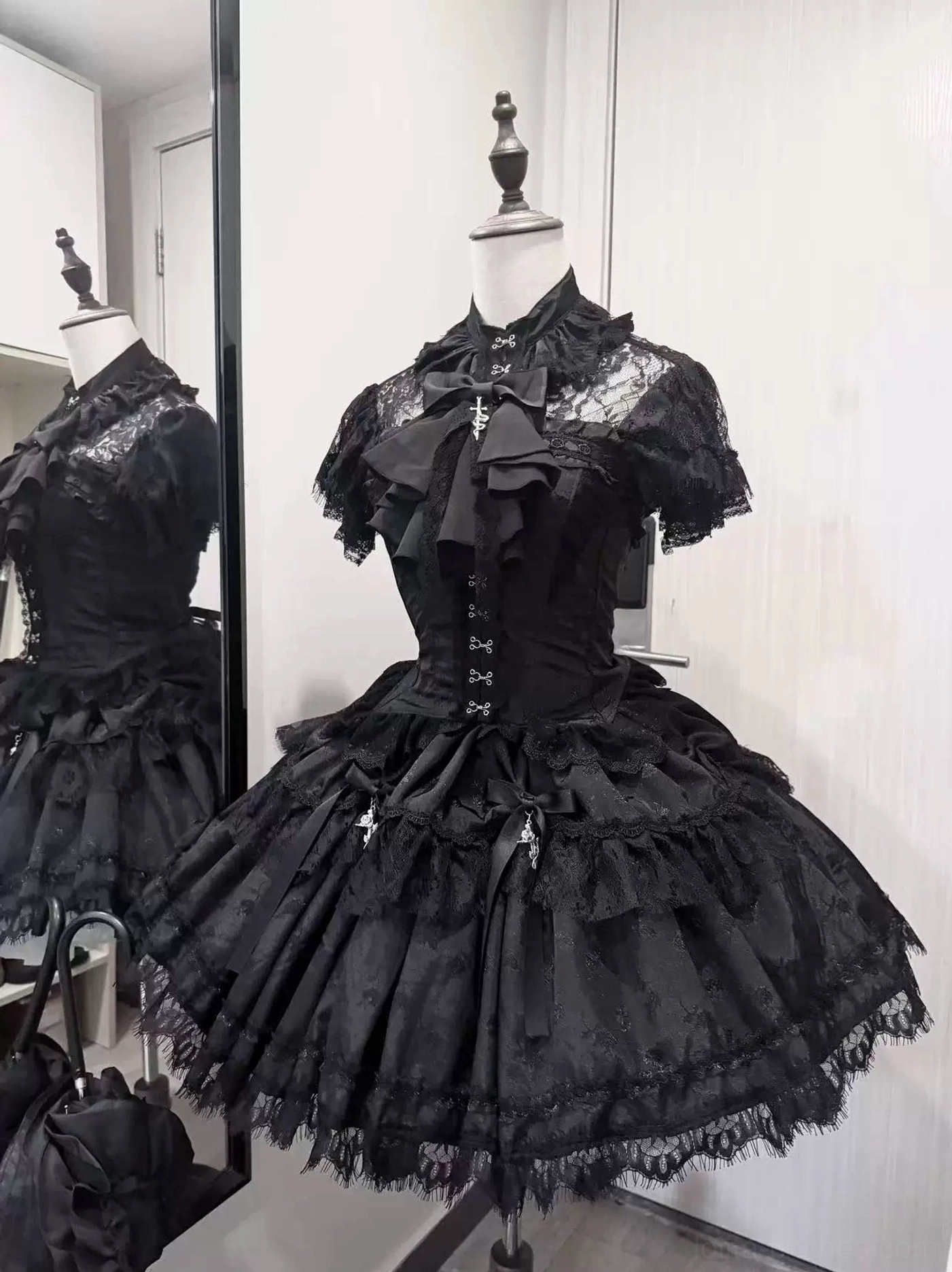 Black gothic lolita corset with short lace sleeves and layered mesh skirt featuring cross pendants, front three-quarter view with side mirror reflection, studio mannequin display