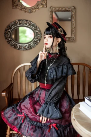 Female model wearing gothic lolita outfit with cross pearl necklace in three-quarter view featuring black ruffled blouse wine-red tiered skirt black fedora hat indoor vintage cafe styling
