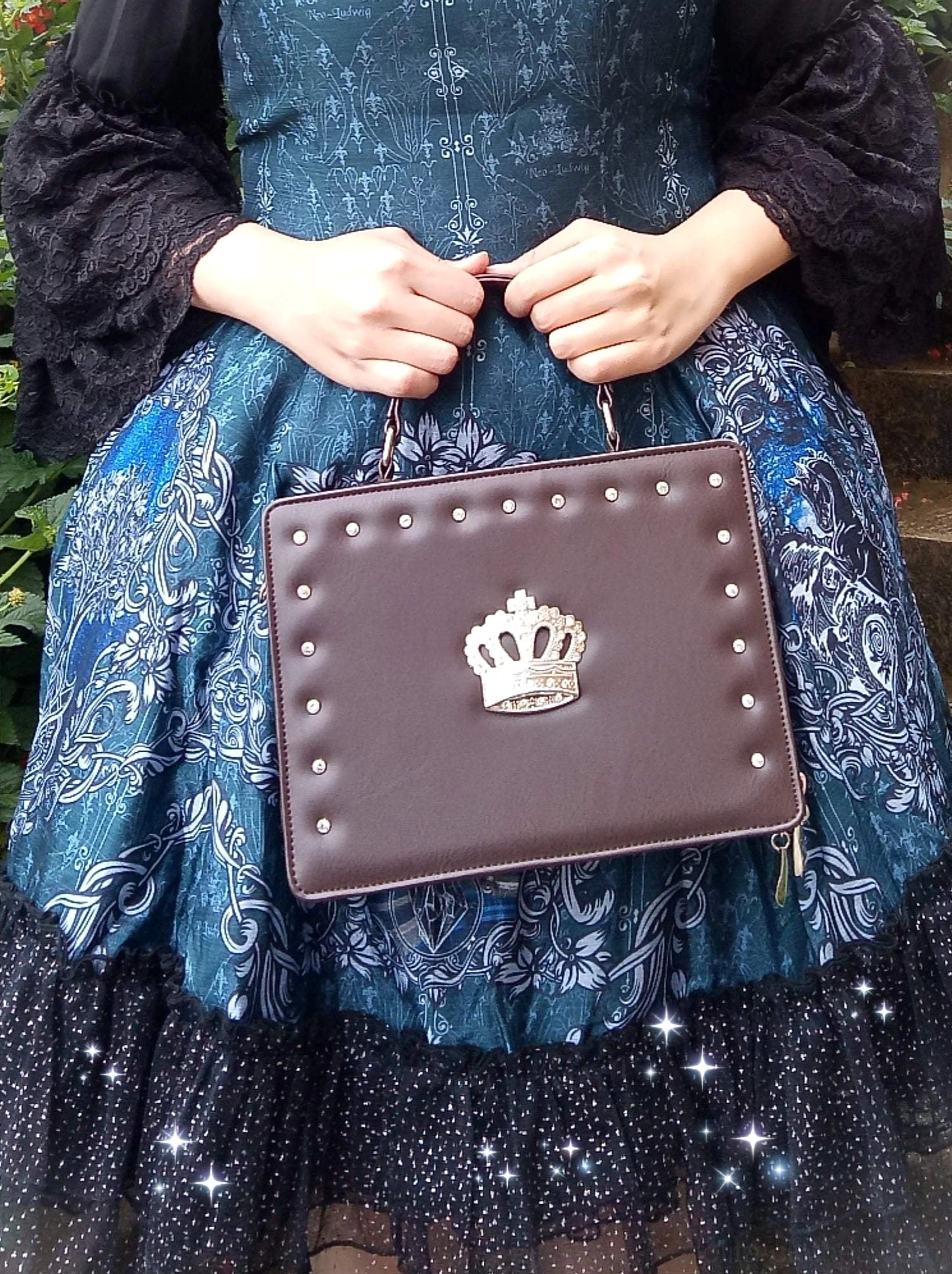 model holding dark brown gothic lolita crown handbag front close-up shot dark brown pu leather studded edges central silver crown top handle indoor blue patterned lolita dress