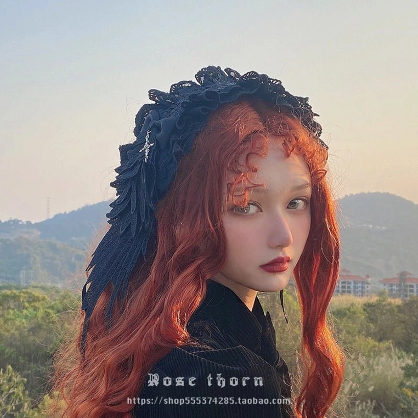 Gothic lolita devil wings headband three-quarter side profile on model with long curly reddish hair, outdoor mountain backdrop, silver cross charm detail visible
