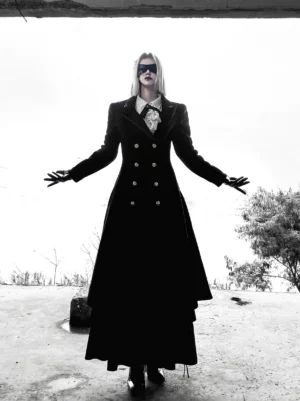 Front full-body view of gothic lolita double-breasted black coat worn by silver-haired model with blindfold and white bow tie in barren outdoor field, showcasing tailored waist silhouette and metal button details