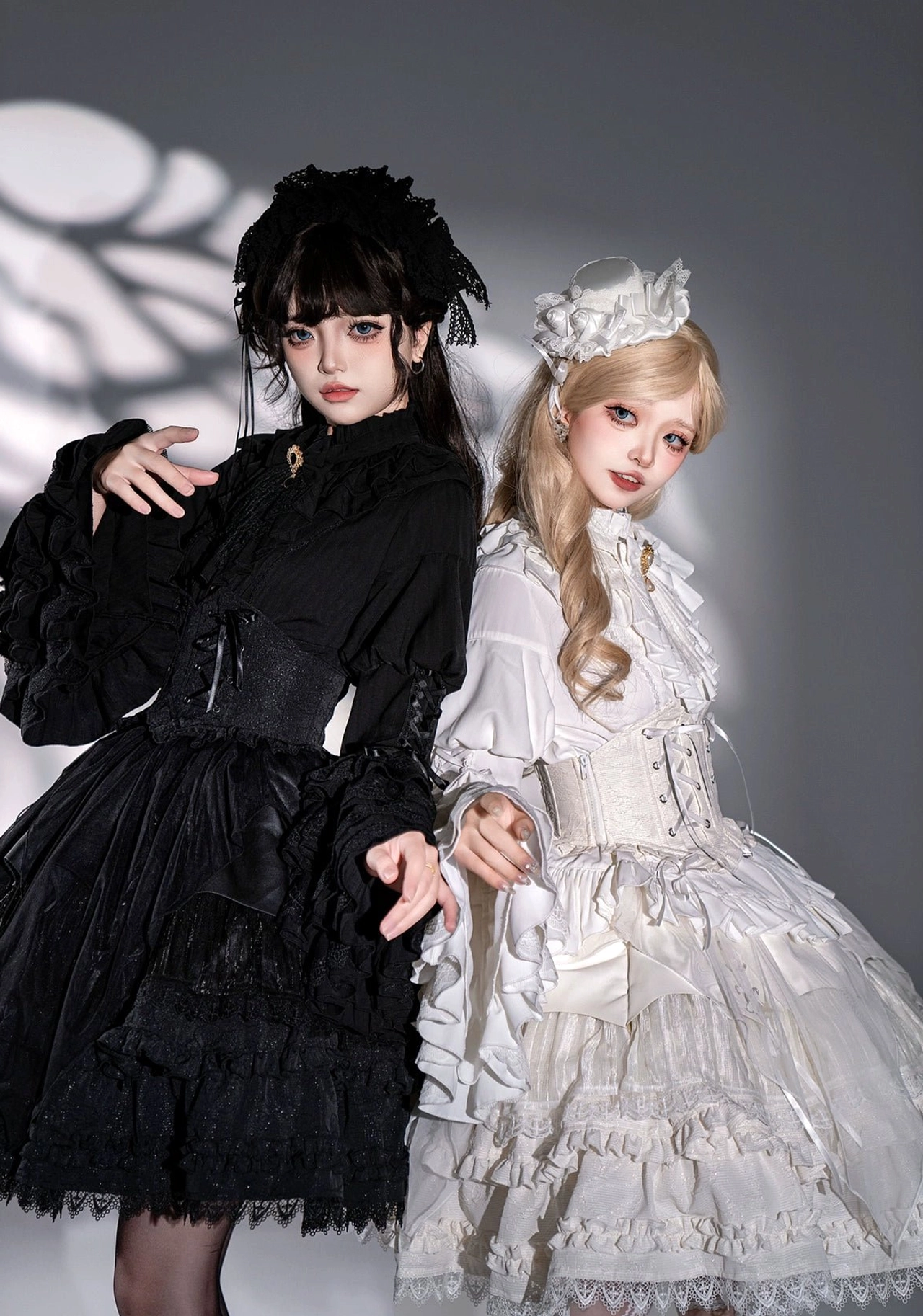 Two models in gothic lolita dresses close side view showing black high-neck long-sleeve dress and white princess dress with tiered ruffles corset waist and hair accessories