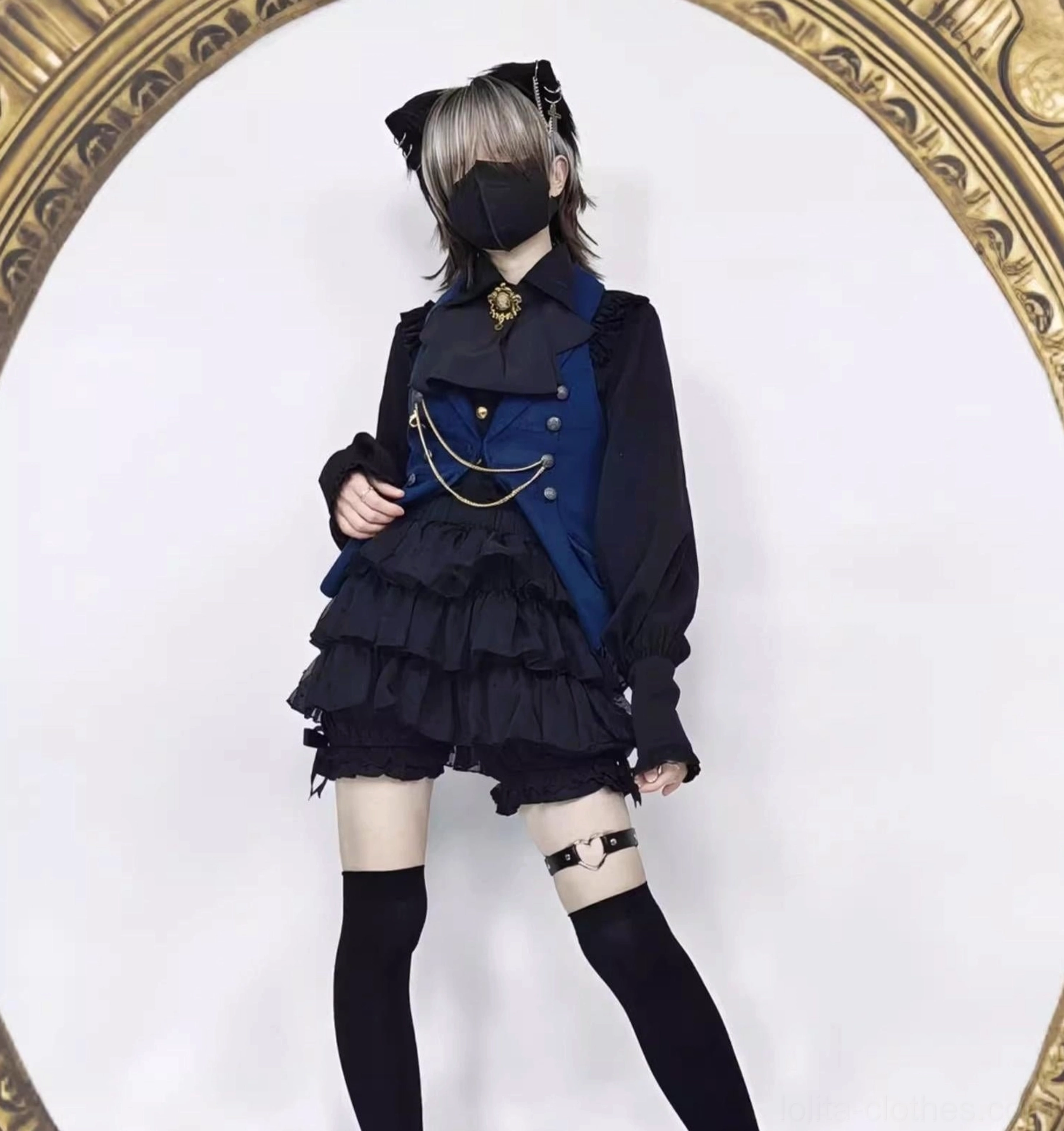 Male lolita model in gothic prince style coordinate featuring black tiered ruffle shorts gold chain vest accessory single leg garter and formal gothic aesthetic styling pose