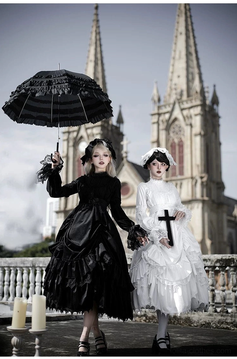 Black and white velvet gothic lolita OP dresses on double models at eye level showing tiered ruffled skirts, cross-adorned collar straps and bow tie collars with parasol and cross props